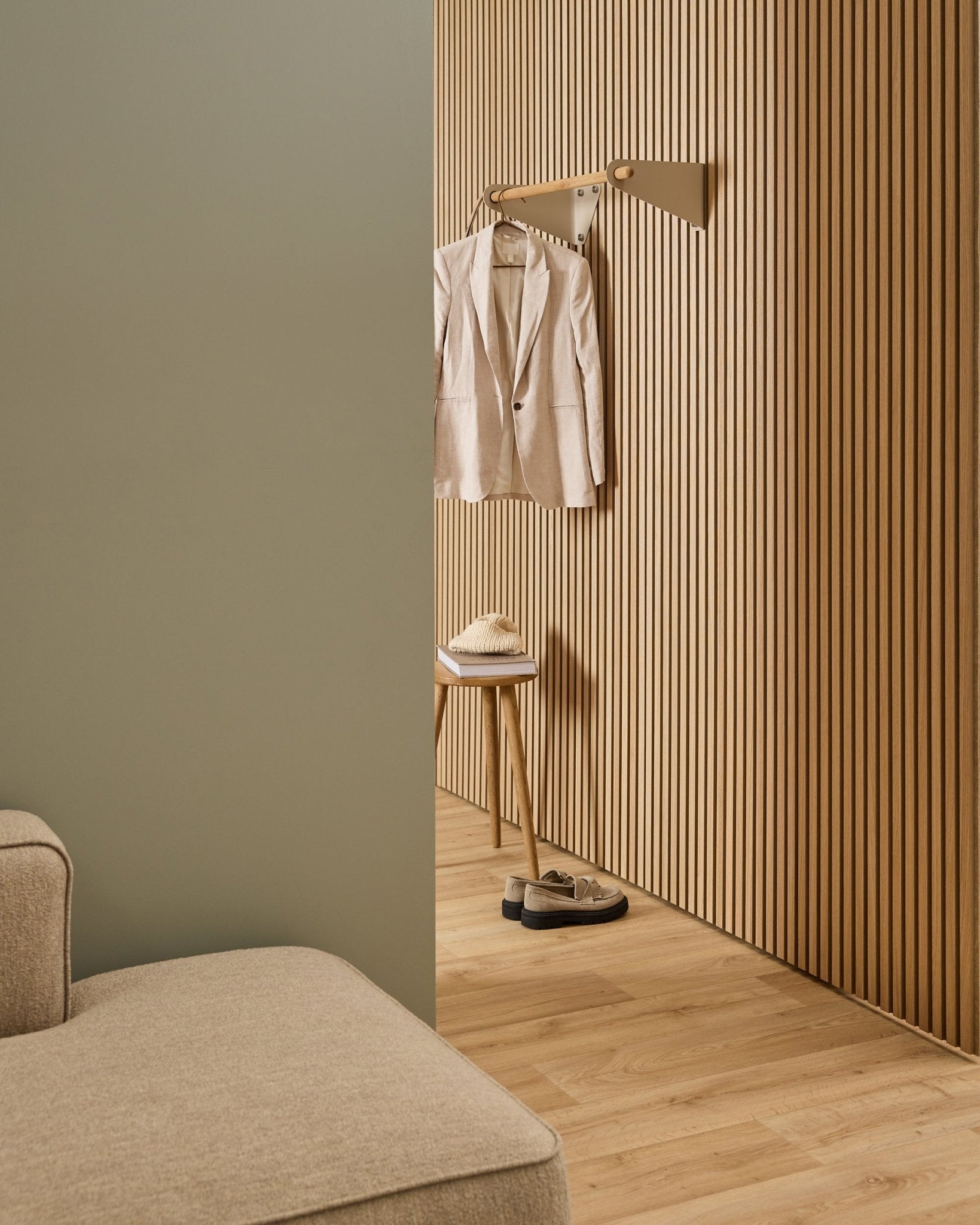 Minimalistischer Raum mit beige Sofa, Akupanel Akustik-Lamellenwand, WoodUpp Linn Garderobenstange in beige (75,9 cm), Hocker mit Hut und Schuhen auf hellem Holzboden.